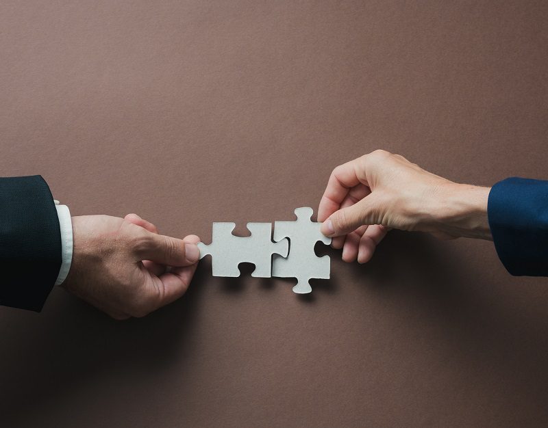 Teamwork and partnership conceptual image - hands of a businessman and businesswoman joining blank matching puzzle pieces over brown background.