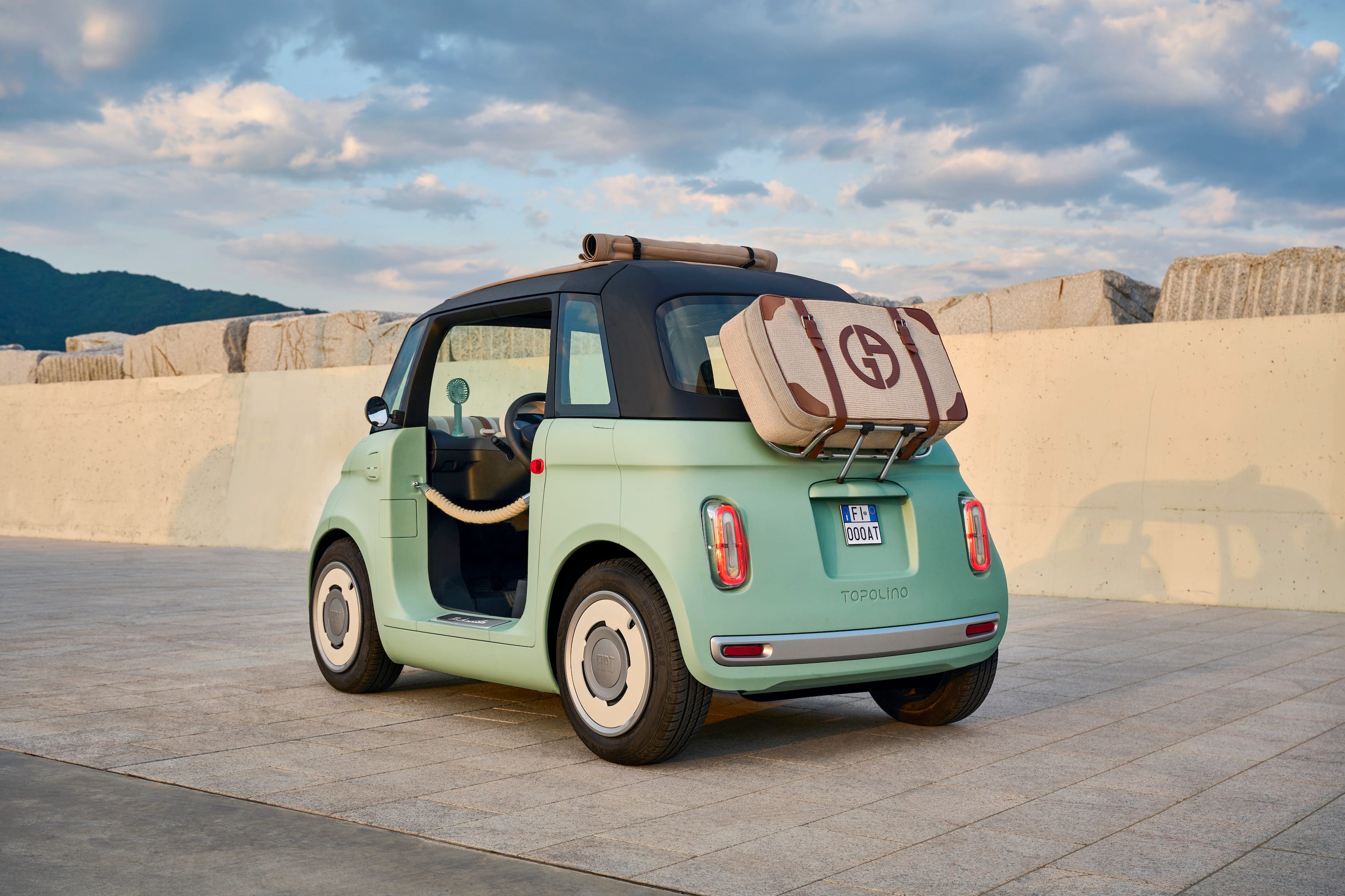 A teal Fiat Topolino Dolcevita microcar with blue skies and clouds in the background.