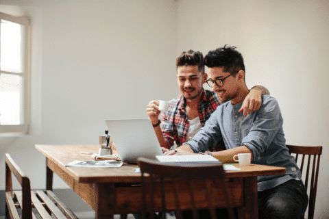 Young guy with cup of fresh coffee hugging boyfriend browsing laptop while sitting at table during breakfast at home