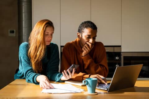 Serious room-mates paying utilities bills at home sitting in the kitchen using laptop and phone. Internet connection to pay expenses