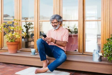 Senior greyish bearded man barefoot wearing a stylish salmon polo shirt and blue jeans using smartphone in a chill city terrace