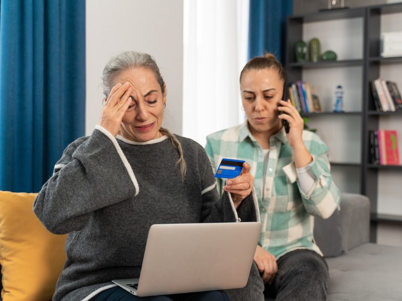 A woman sits with her laptop and credit card in one hand and is holding her head. Another woman is behind her talking on the phone