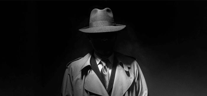 A man wearing a fedora and trench coat standing mysteriously in the shadows