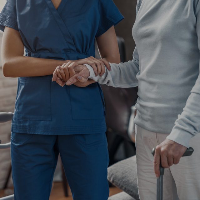 An aide in scrubs helping an older woman walk.