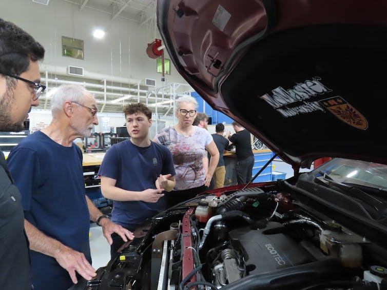 a group of older people and students stand around the open hood of a car