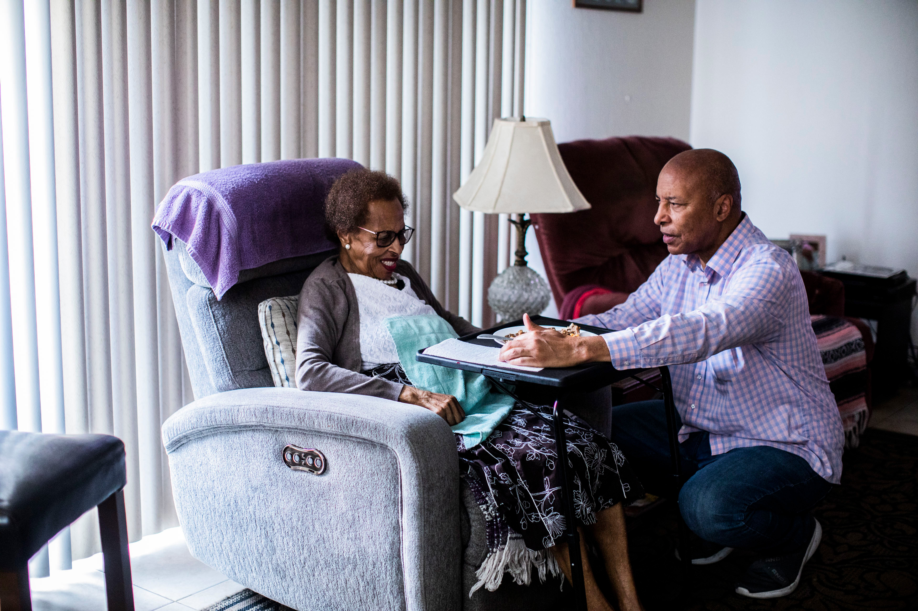 A photo of an elderly woman's son taking care of her while she sits in a chair.