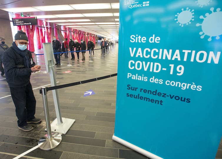 A sign in the foreground for a COVID-19 vaccination clinic, with people lined up in the background.