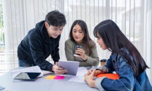 a group of three students looking at a tablet or laptop in the middle of a table littered with class papers and notes