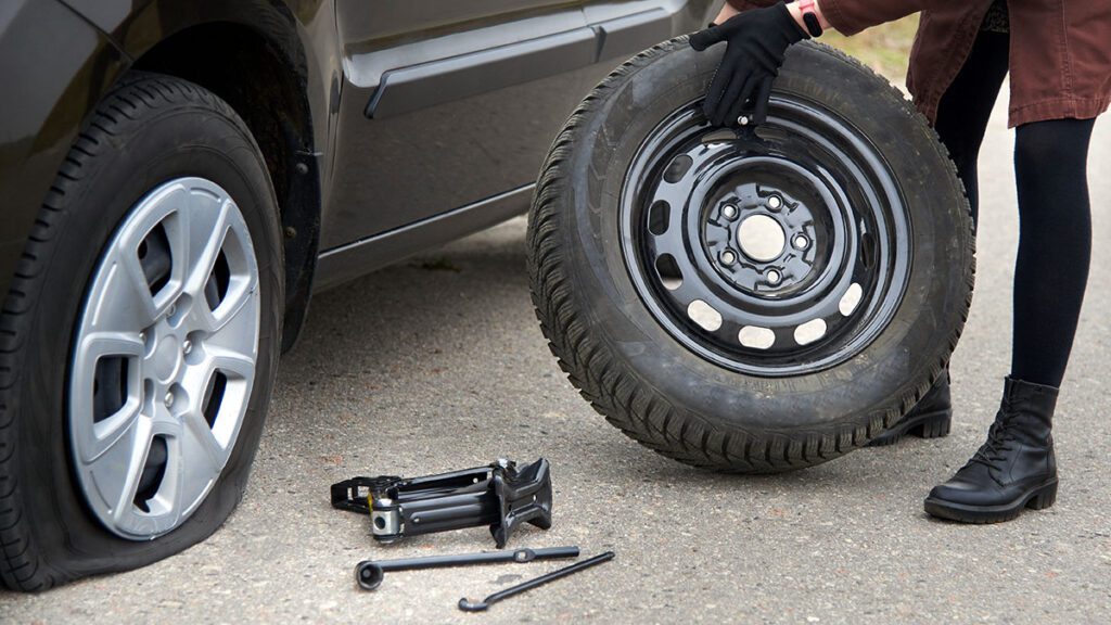 A young woman rolls spare tire near her car with a flat tire, trouble on the road.