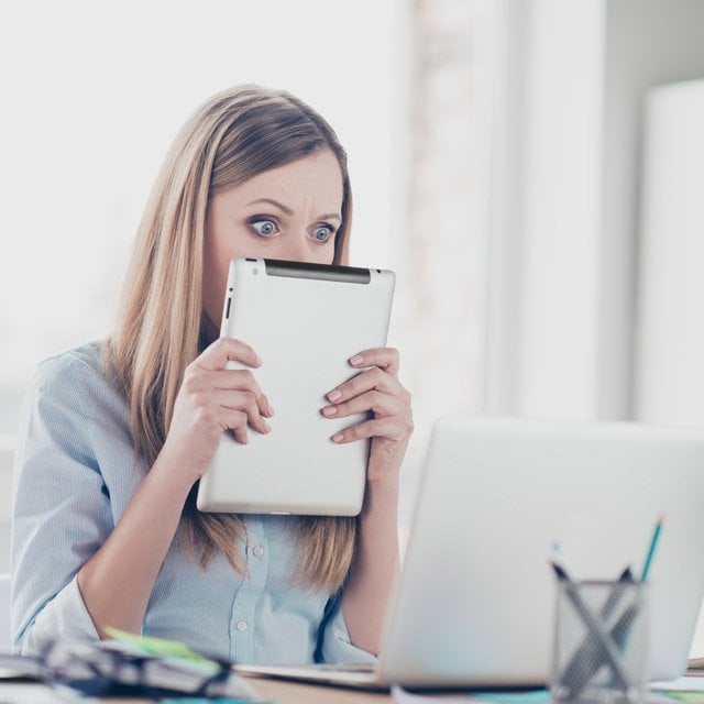 A Woman covering half face with tablet looking with wide open eyes at laptop