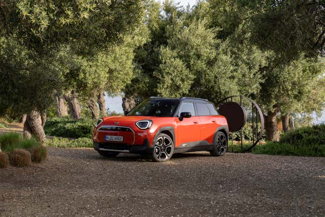 An orange Aceman SE  parked on gravel in front of a sculpture and trees