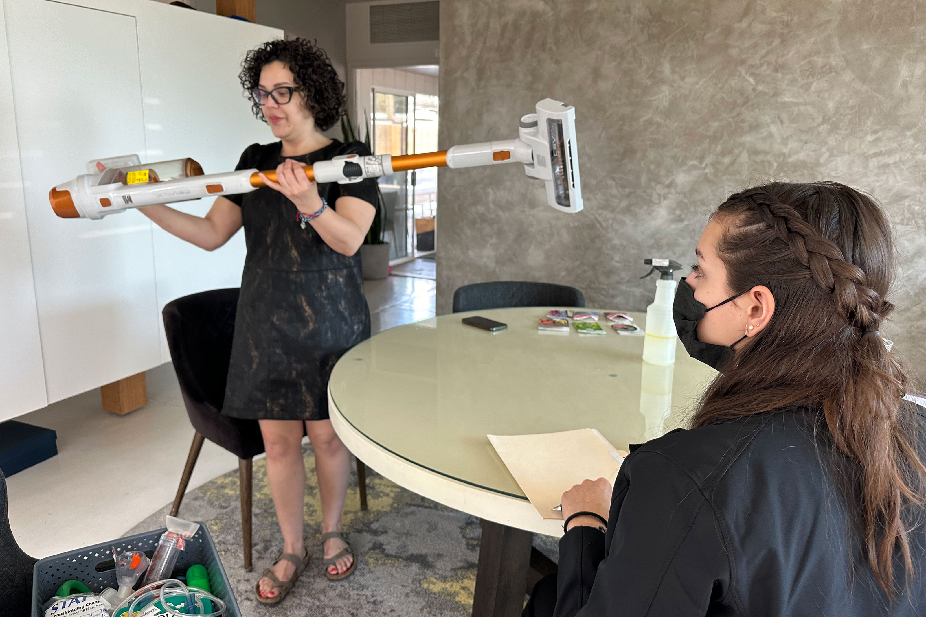 A photo of a mother holding up a vacuum cleaner while a community health worker is sitting at a table.