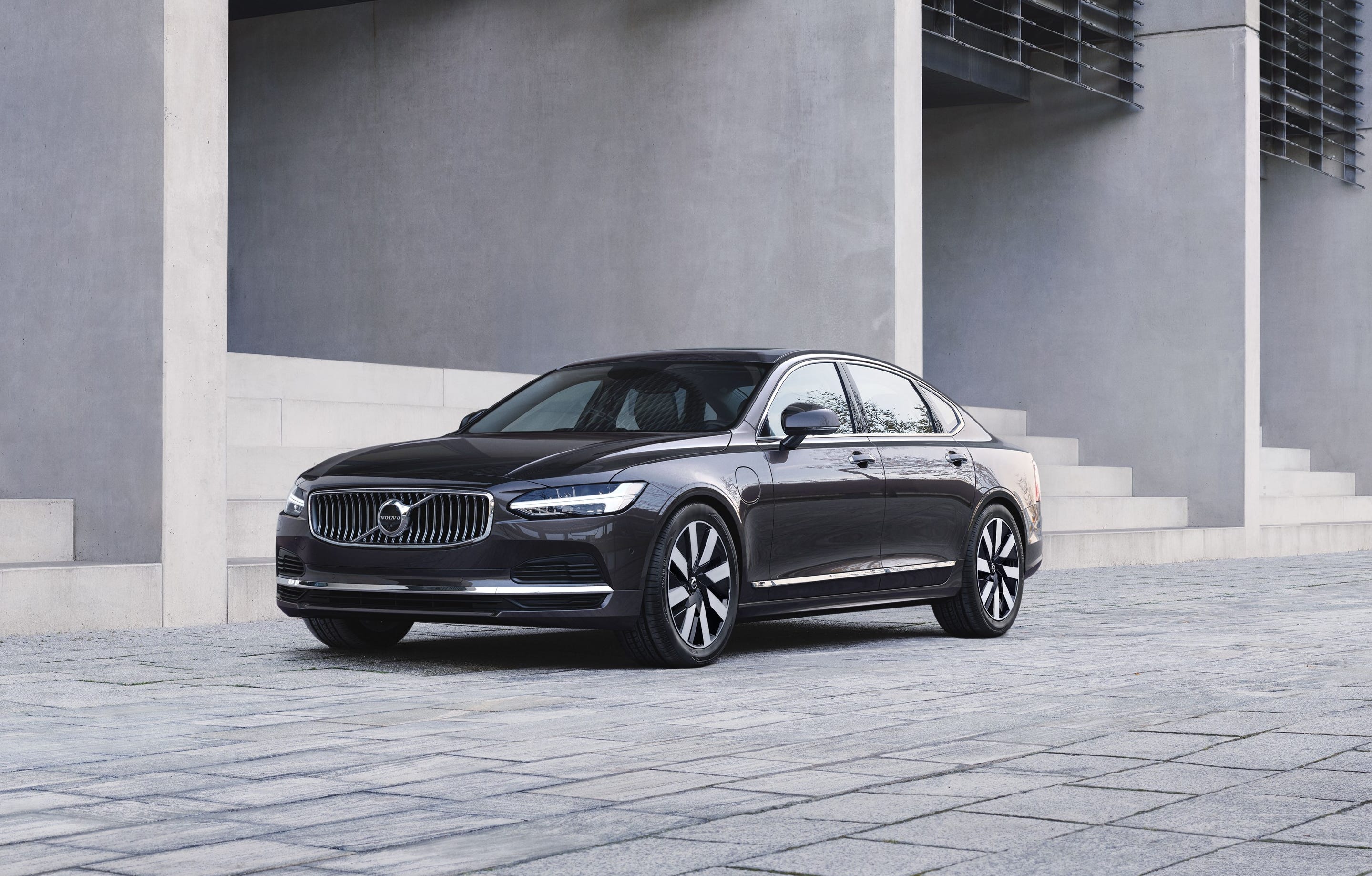 A grey Volvo S90 Recharge T8 AWD sedan is parked in front of a concrete building.