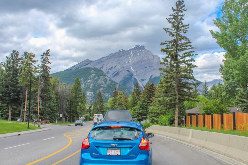 Rear view of a blue car on a tree-lined road, heading towards distant mountains under a partly cloudy sky. Taken at highway from Calgary to Banff National Park, Alberta, Canada on June 2019.