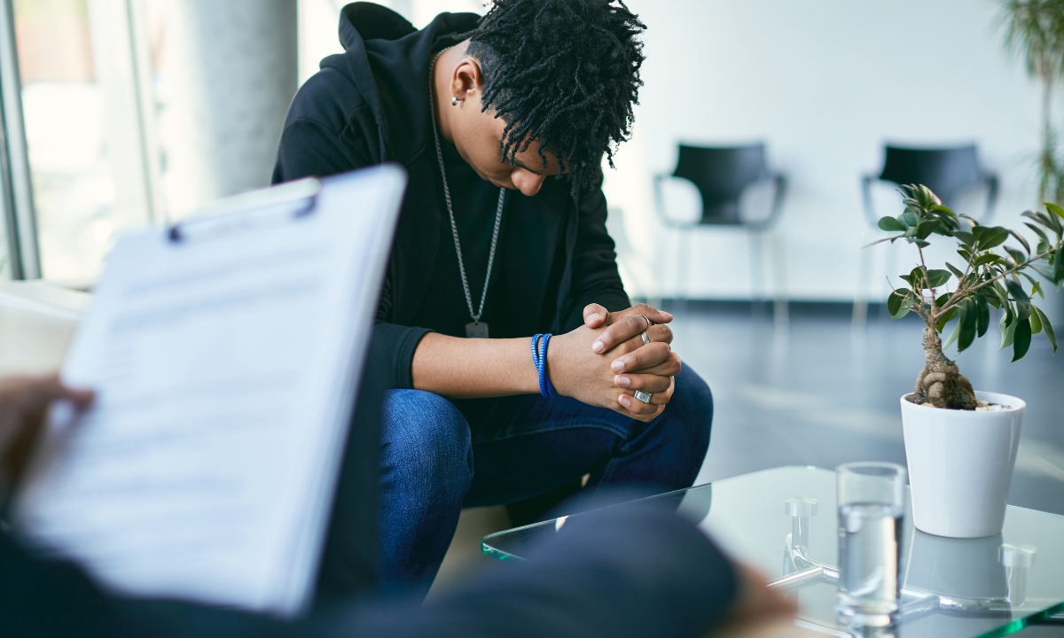 a man in a therapy session, his hands are clasped and his knees are spread as he stares at the ground