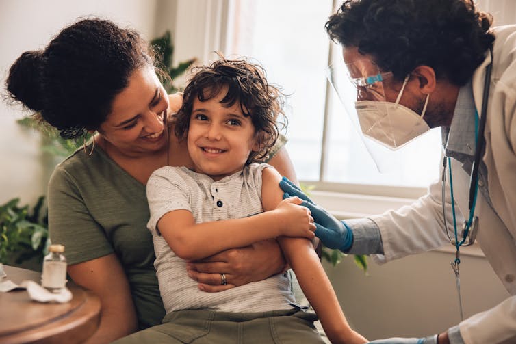 A mother holding a smiling child in her lap. The doctor attends to the child's upper arm following a vaccination.