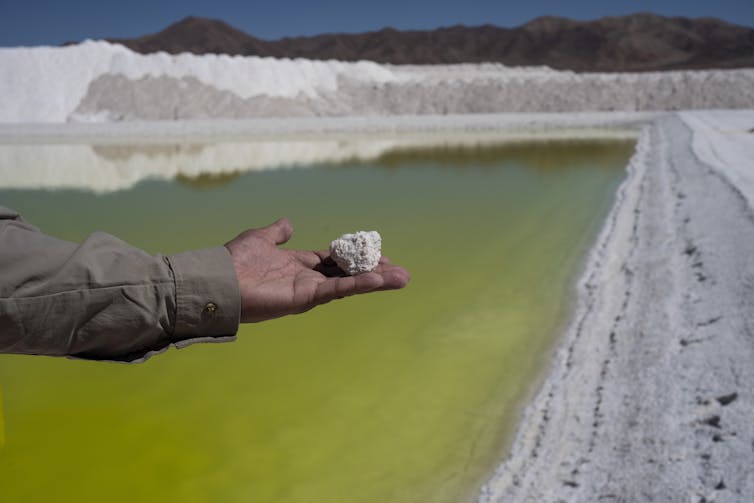 An outstretched hand holds a chunk of salt against the backdrop of a pool of water.