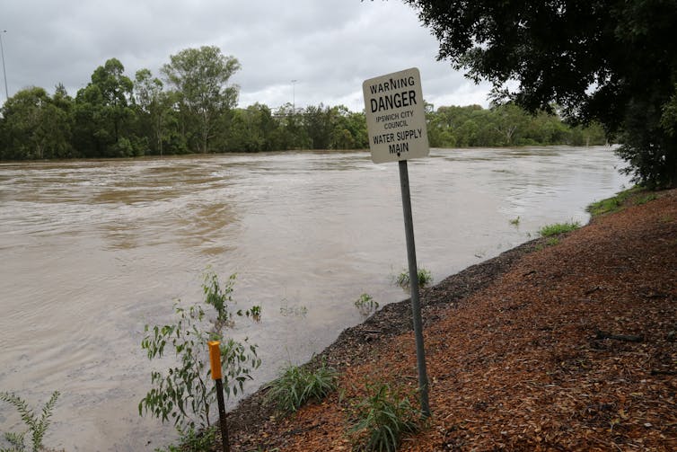 Floodwaters flow past a warning sign about a water supply main in Ipswich, Queensland