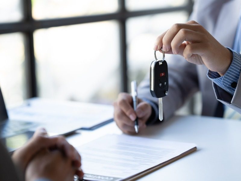 To the right, a car salesperson holds a set of keys above a contract, which sits on the desk. Across from the salesperson is the buyer.