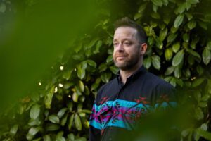 A photo of a man standing outside for a portrait by some shrubbery.