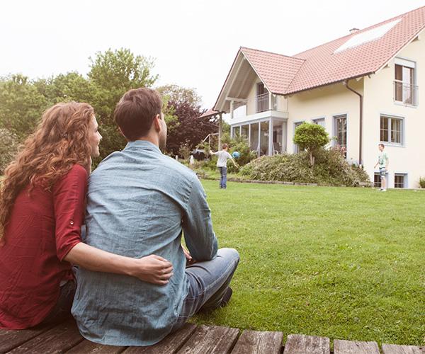 Two people sitting on a bench looking at their home. A younger child is playing on the lawn.