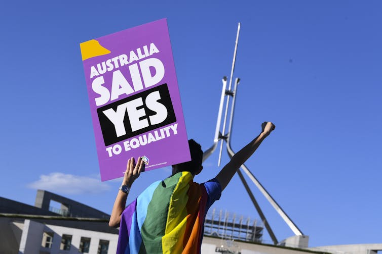 Person with LGBTIQ+ flag wrapped over shoulder holding 'Australia said yes to equality' placard, fist in air