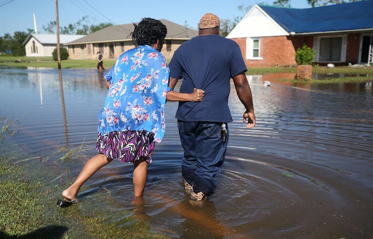 A woman holds onto the back of a man's shirt as they step through calf-deep water cover a road. Brick one-story homes are on the other side.