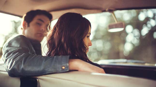 A view from behind a parked young man and woman as visible from the back seat of their classic 1960s car as they have a disagreement or relationship difficulties.