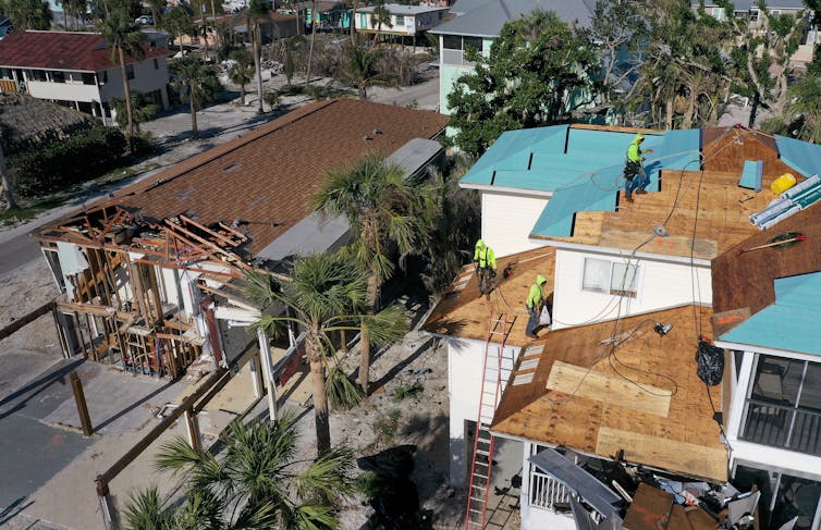 Workmen repairing the roof of a damaged house, surrounded by other homes with roof damage.