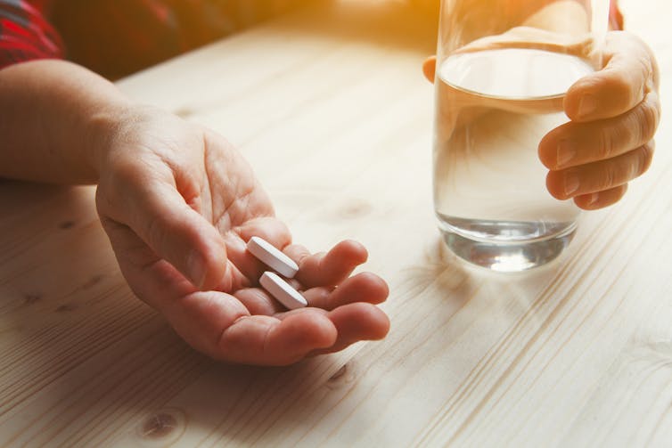 A hand holding two white tablets, with another hand holding a glass of water, on a table.