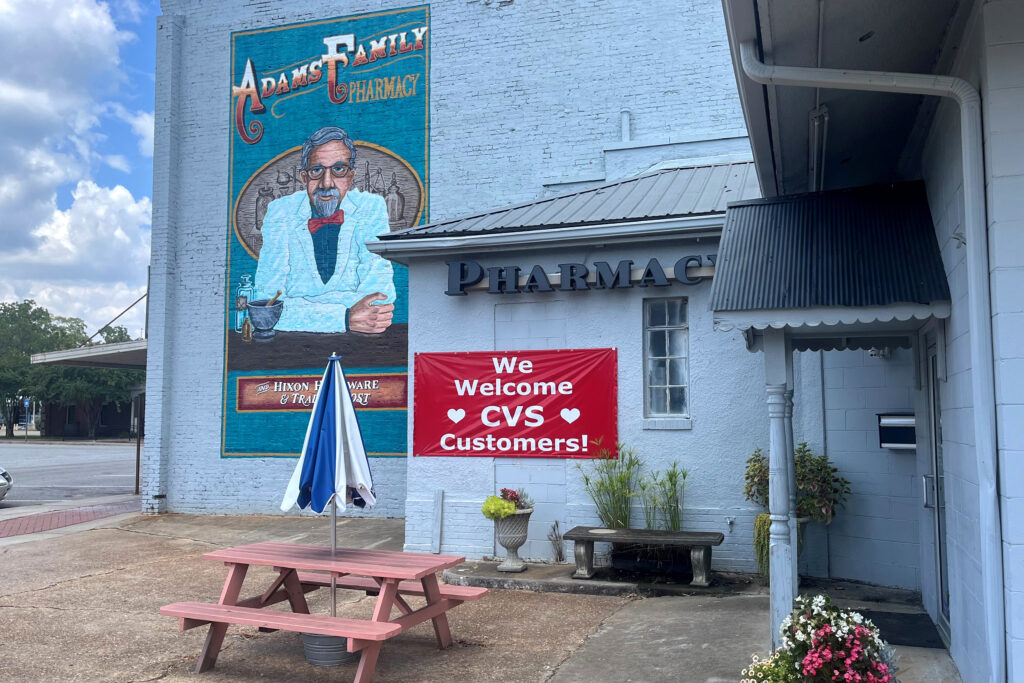 A photograph of the exterior of the Adams Family Pharmacy on a sunny day. There is a red sign out front that reads: "We Welcome CVS Customers!"