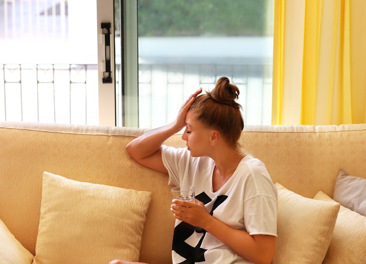 A young woman holds sitting on a yellow couch holds her head and a glass of water.