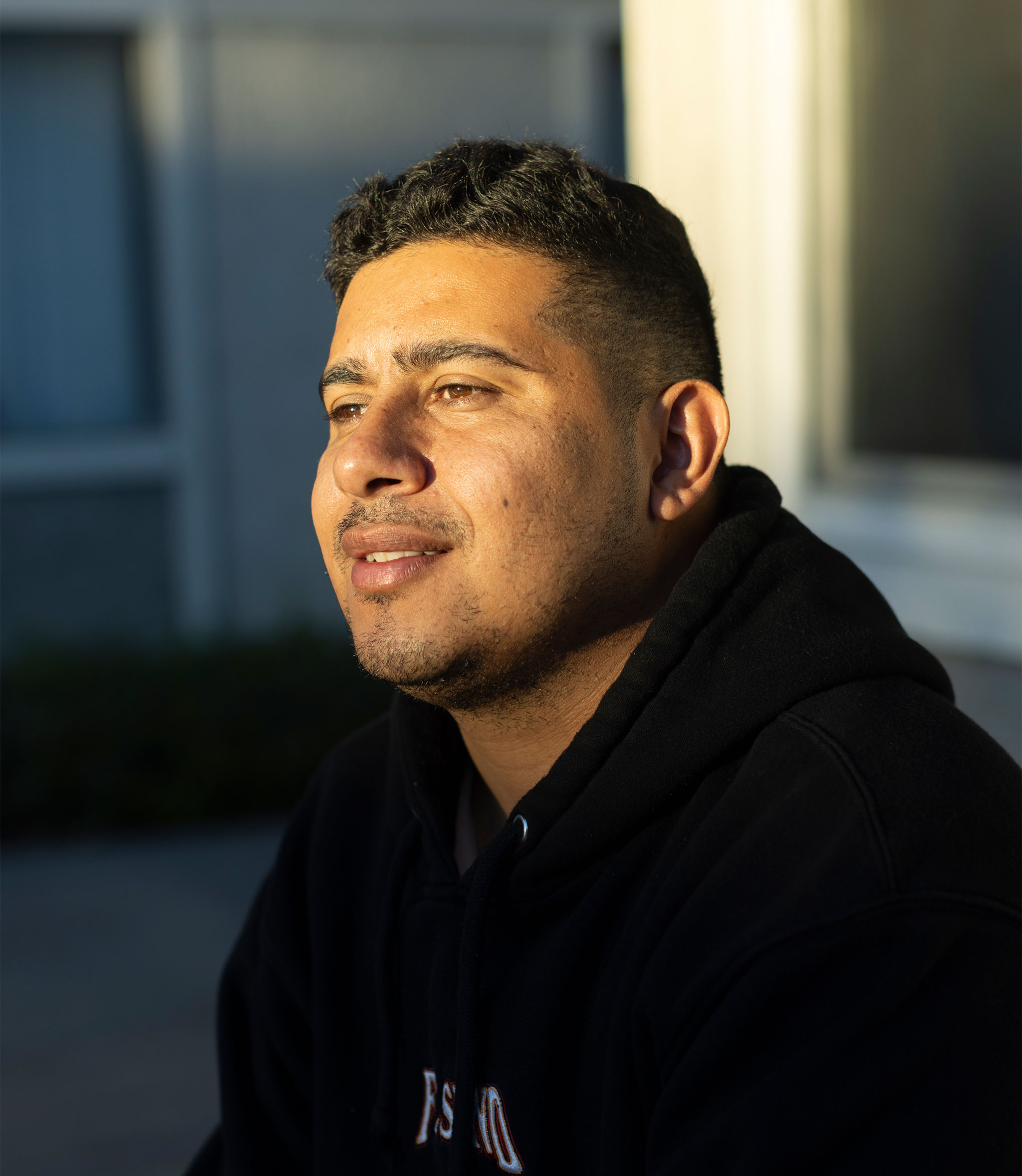 A man in a black hoodie smiles slightly and looks to his right as the sun shines on his face.