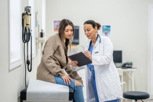 Teenager at a Medical Appointment A young female teenager sits up on an exam table during a routine medical appointment with her doctor. She is dressed casually and leaning in to look at her test results as her doctor shares them on a tablet. health insurance stock pictures, royalty-free photos & images