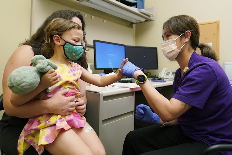 A woman holds a young girl in a pink dress, who is wearing a mask and holding a stuffed animal, as she fist-bumps a nurse in a purple shirt.