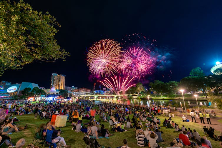 A crowd of people on the grass watching pink, blue and orange fireworks in the night sky.