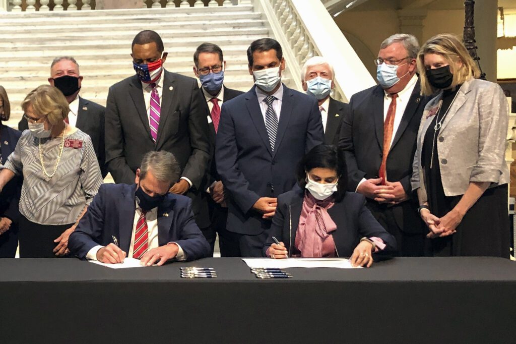 Georgia Gov. Brian Kemp (left) and Centers for Medicare & Medicaid Services Administrator Seema Verma (right) sit at a table facing the camera while they sign papers. Nine other individuals in business attire stand in a row behind them. Everyone is wearing face masks.