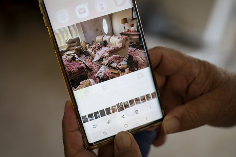 A cell phone displays a photo of a home interior with severe hurricane damage.