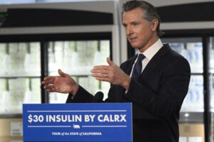 California governor Gavin Newsom stands behind a blue and white sign that reads "$30 insulin by CalRx"