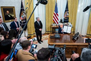 A photo of President Trump holding up a signed executive order while surrounded by news cameras and microphones.