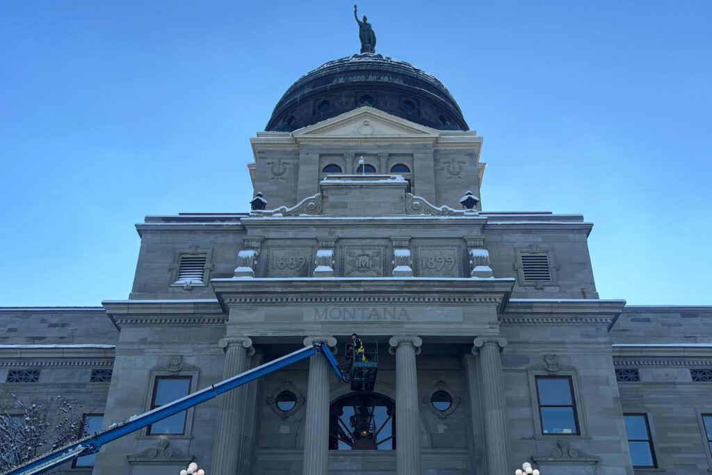 A photo of the exterior of the Montana Capitol. A worker in a construction vest is seen in front of the entrance in a cherry picker.